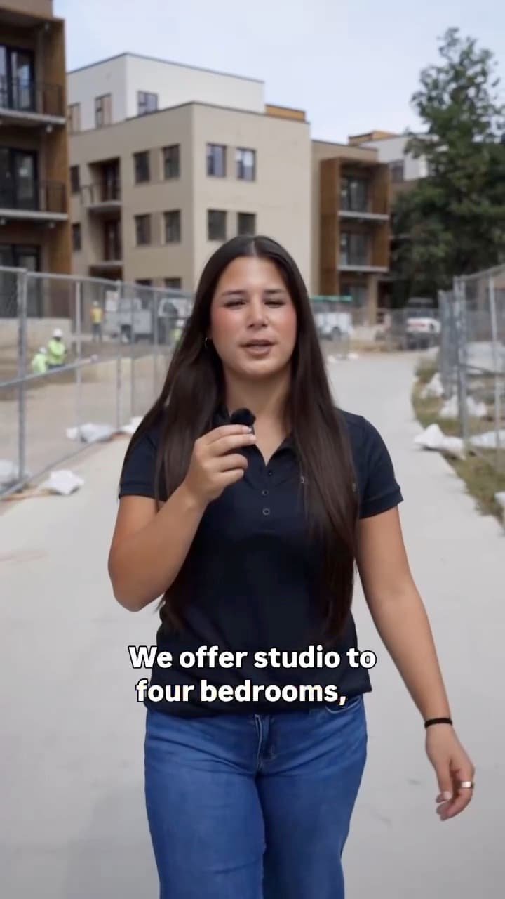 A host speaking to camera on a path beside new apartment buildings